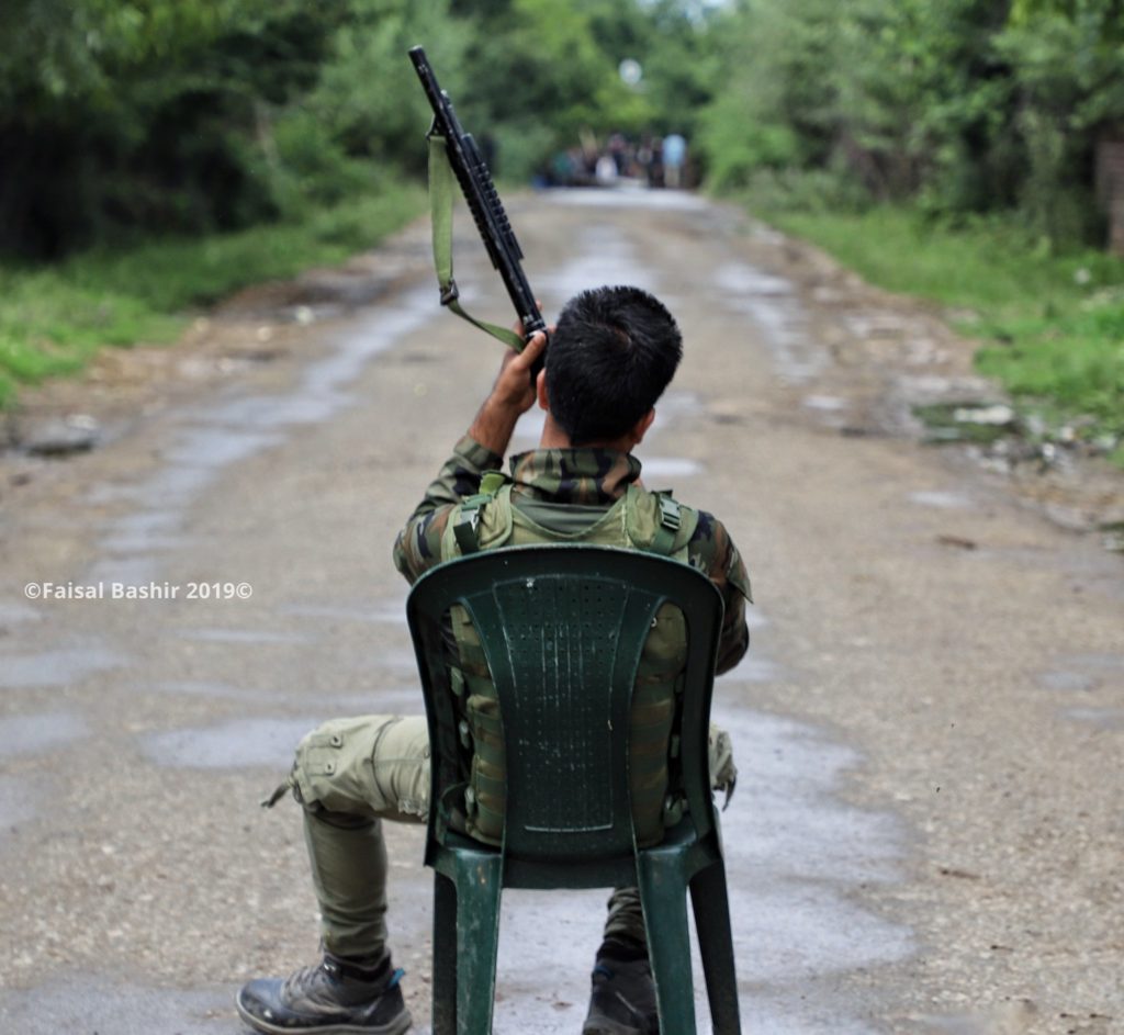 indian army personnel sitting on chair as he shows his weapon gun towards kashmiri protesters in Shopian